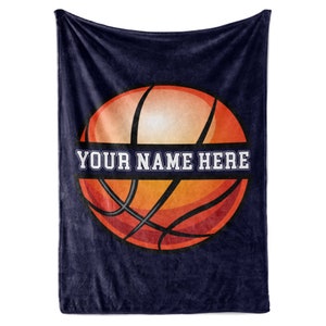 May include: A navy blue fleece blanket featuring a basketball design. The basketball is orange and brown with black lines, and the text "YOUR NAME HERE" is in white. The blanket is a soft, cozy item.