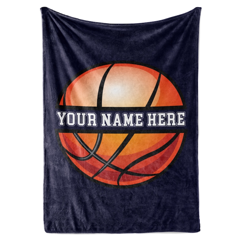 May include: A navy blue fleece blanket featuring a basketball design. The basketball is orange and brown with black lines, and the text "YOUR NAME HERE" is in white. The blanket is a soft, cozy item.