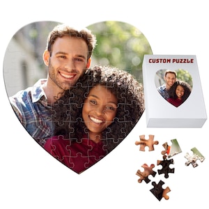 May include: A heart-shaped custom puzzle featuring a smiling couple. The puzzle is partially assembled, with a white box displaying the same image. The box is labeled "CUSTOM PUZZLE". Several puzzle pieces are scattered nearby.