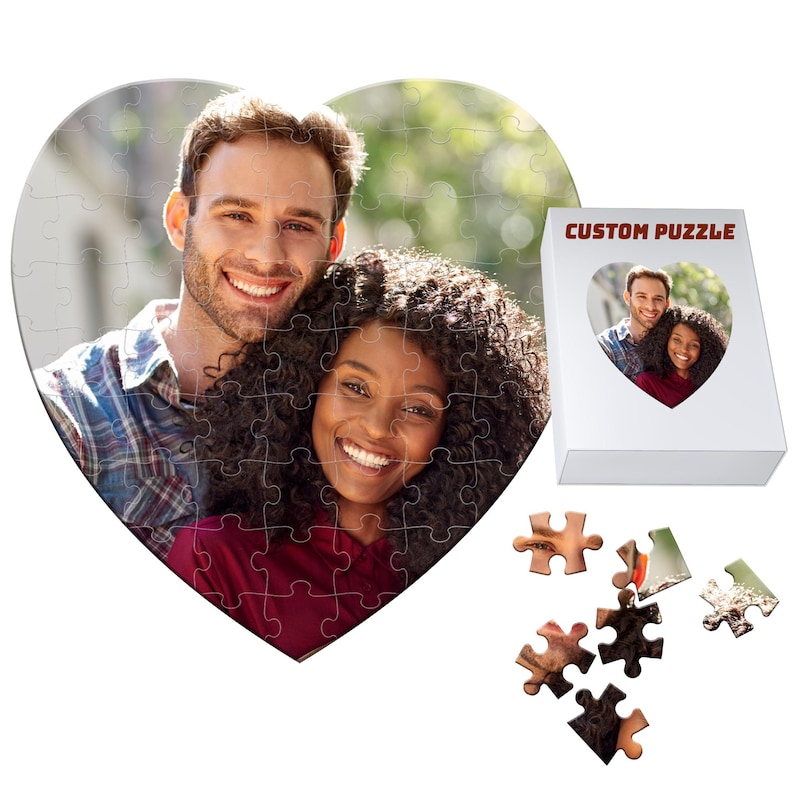 May include: A heart-shaped custom puzzle featuring a smiling couple. The puzzle is partially assembled, with a white box displaying the same image. The box is labeled "CUSTOM PUZZLE". Several puzzle pieces are scattered nearby.