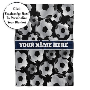 May include: A black blanket featuring a pattern of soccer balls in shades of gray and white. A navy blue banner across the blanket reads "YOUR NAME HERE" in white. The blanket also has the text "Click Customize Now To Personalize Your Blanket".