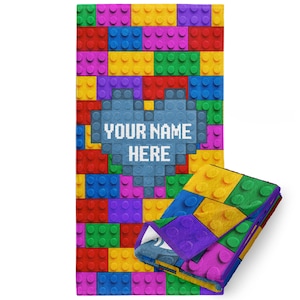 May include: A colorful beach towel with a pattern of colorful Lego bricks. The towel has a blue heart in the center with the text "YOUR NAME HERE" in white.