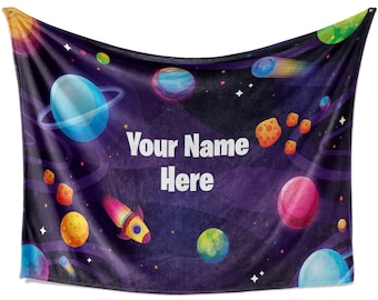 Puede incluir: Una manta de forro polar morada con temática espacial. Presenta planetas, estrellas, una nave espacial y el texto "Your Name Here".