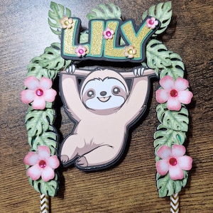 May include: A cake topper featuring a sloth holding a sign that says "LILY" with green leaves and pink flowers on either side. The sloth is brown and has a cute face. The sign is gold with glitter.