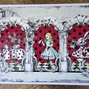 May include: A white metal box with a vintage design featuring Alice in Wonderland characters. The box has three arched sections with a red and white polka dot background. The characters include Alice, the Queen of Hearts, and the White Rabbit. The box has a clock face with Roman numerals on the left side.