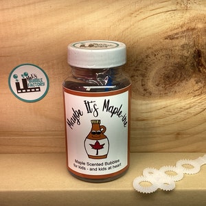 May include: A clear bottle of maple scented bubble solution with a label that says "Maybe It's Maple-y" and features a cartoon maple syrup bottle with a Canadian flag. The bottle is sitting on a wooden surface next to a white plastic bubble wand.