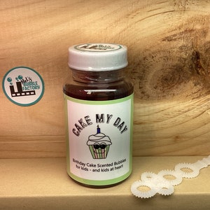 May include: A bottle of birthday cake scented bubble solution with a green label that says "Cake My Day" and a cartoon cupcake with a candle on top. The bottle is on a wooden surface with a white bubble wand.