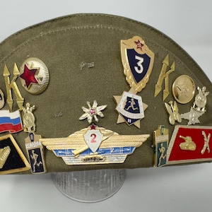 May include: A green military style hat with a variety of pins and badges attached. The pins include a red star, a tank, a plane, and a number "2" with a red star above it.