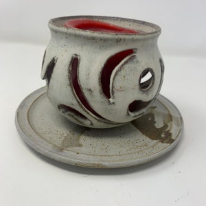 Peut inclure: Bougie blanche en céramique avec une bougie rouge à l'intérieur. Le bougeoir a un motif décoratif de volutes découpées. Il repose sur une soucoupe en céramique blanche assortie.