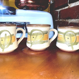 May include: Three ceramic mugs with brown, green, and orange abstract designs. The mugs have a cream-colored background and are all slightly different.