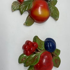 May include: Two vintage wall decorations featuring red apples, green leaves, and blue and purple plums. The apples are painted in a realistic style with red, yellow, and orange hues. The leaves are green and the plums are blue and purple.