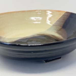 May include: A ceramic bowl with a black, brown, and cream glaze. The bowl has a wide, shallow shape and a slightly flared rim.