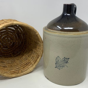 May include: A vintage stoneware crock with a brown lid and a blue and white maple leaf design. The crock has the text "Western Vinegar Co." printed on it. The crock is next to a woven basket.