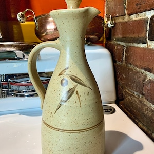 May include: A ceramic olive oil bottle with a brown and tan speckled glaze. The bottle has a brown lid and a brown handle. The bottle is decorated with a brown olive branch design.