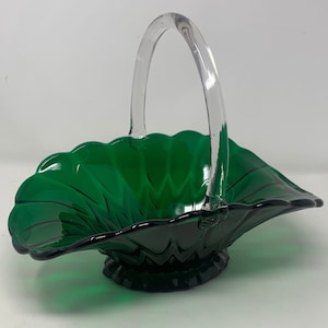 Può includere: Un cestino vintage in vetro verde con manico in vetro trasparente. Il cestino ha un bordo smerlato e una base a piedistallo. Il vetro è di un colore verde smeraldo intenso e il manico si incurva con grazia sopra il cestino.