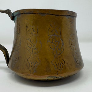 May include: A brass cup with a handle, featuring intricate engraved designs. The cup has a rounded bottom and a flared top.