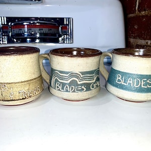 May include: Three ceramic mugs with brown rims and handles. The mugs have different designs. The first mug has the text "BLADES & TRICKS" in brown. The second mug has the text "BLADES GALLERY" in blue. The third mug has the text "BLADES AND" in blue.