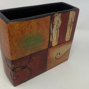 May include: A rectangular ceramic vase with a geometric design in shades of red, brown, yellow, and green. The vase has a black rim and a textured surface.