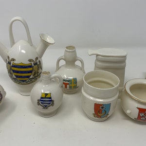 Peut inclure: Ensemble de huit petits pots en céramique blanche avec des garnitures dorées et des armoiries colorées. Les pots sont de formes et de tailles variées, notamment une théière, un pichet et un pot rond.