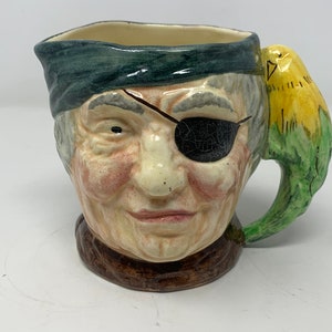 May include: A ceramic pitcher shaped like a pirate's head with a green eye patch and a yellow bird perched on the handle.