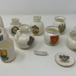 May include: A collection of 10 miniature ceramic teacups and a teapot, each decorated with a different coat of arms. The cups are white with gold trim and feature colorful crests.