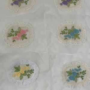 May include: Six white fabric squares with embroidered floral designs in various colors. Each square features a circular lace trim around the embroidered design.
