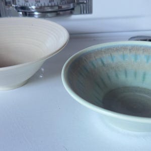 May include: Two ceramic bowls, one white and one with a light green and brown glaze. The bowls are both round and have a slight curve to them.