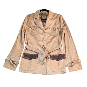 Puede incluir: Chaqueta color beige con cuello, cierre de botones y cinturón a juego. La chaqueta presenta dos bolsillos en el pecho y dos bolsillos inferiores con detalles de cuero marrón oscuro. Las mangas tienen un detalle anudado en los puños.