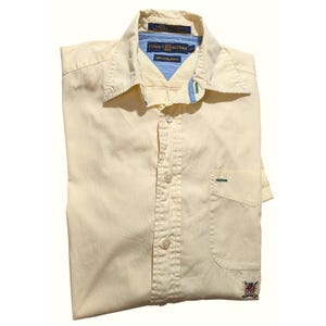 Puede incluir: Camisa abotonada amarilla clara doblada, con rayas verticales. La camisa presenta un bolsillo en el pecho con un pequeño acento verde y un logotipo en la parte inferior. El cuello y el forro interior son de un azul claro contrastante. La marca "Tommy Hilfiger" es visible.