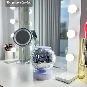 K&ouml;nnte beinhalten: Ein silberner Discokugel-f&ouml;rmiger &auml;therischer &Ouml;ldiffusor mit wei&szlig;em Sockel. Der Diffusor ist mit blauem Licht beleuchtet. Der Diffusor steht auf einer wei&szlig;en Oberfl&auml;che mit einem Spiegel und Make-up-Pinseln im Hintergrund.