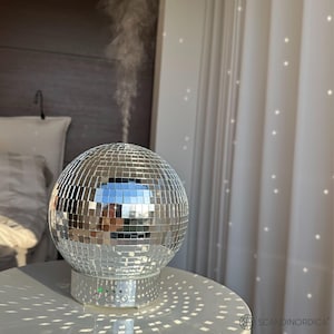May include: A silver disco ball diffuser with a white base and a steam plume rising from the top. The diffuser is on a white table with a silver disco ball pattern.