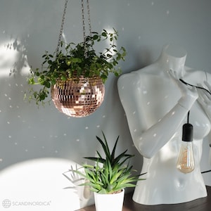 May include: A hanging planter made of a mirrored disco ball with a leafy green plant inside. The planter is hanging from a silver chain. A white mannequin with a light bulb hanging from its chest is in the background.