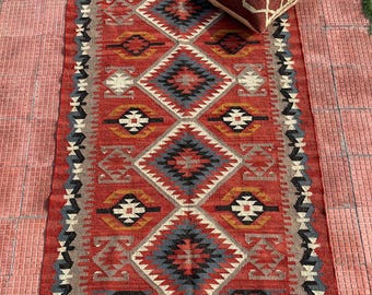 May include: A red, blue, and white patterned rug with a geometric design. The rug has fringe on the edges and is laid out on a red tiled floor.