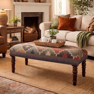May include: A living room scene with a patterned ottoman, dark wooden legs, and a beige sofa with orange and green cushions. A woven tray with a teapot and cups sits on the ottoman. A fireplace and a lamp are also visible.