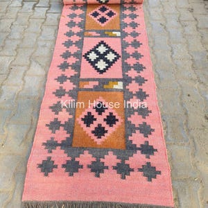 May include: A pink and grey handwoven kilim rug with a geometric pattern. The rug has a fringe on both ends and the text "Kilim House India" is visible in the centre.