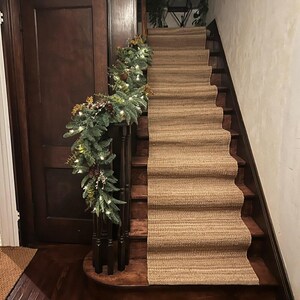 May include: A brown and beige stair runner with a textured woven pattern. A green garland with white lights and pine cones decorates the railing of the staircase.