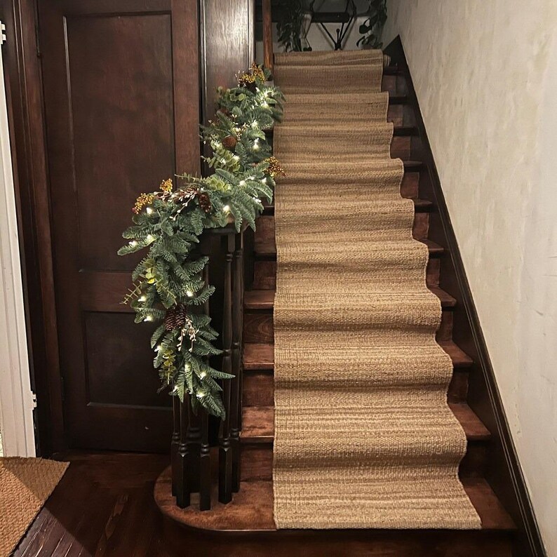 May include: A brown and beige stair runner with a textured woven pattern. A green garland with white lights and pine cones decorates the railing of the staircase.