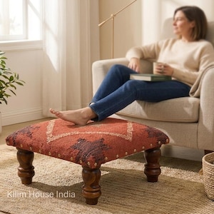 May include: A square, upholstered footstool with a red and brown patterned top and dark wooden legs. The footstool is placed on a woven rug. The image also includes a chair, a plant, and a basket.