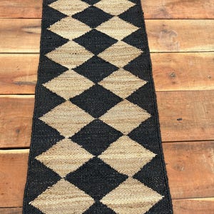 May include: A black and tan diamond-patterned runner rug. The rug is made of woven material and features a repeating pattern of black and tan diamonds. The runner is rectangular and appears to be placed on a wooden surface.