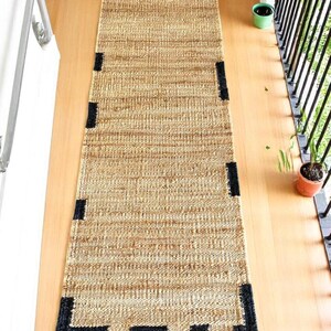 Op de afbeelding: Een rechthoekig jute vloerkleed in natuurlijk bruin met zwarte hoekaccenten. Het kleed is gemaakt van geweven vezels en ligt op een lichtgekleurde houten vloer. Het is een lange, smalle loper.