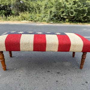 May include: A rectangular bench with a patriotic design. The top features the American flag with red and white stripes and a blue field with white stars. The bench has four wooden legs.