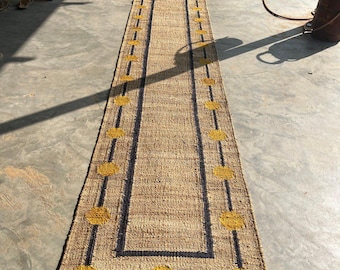 May include: A long, rectangular jute runner rug with a natural tan color. The rug features a dark border and yellow circular accents. The text "KilimHouseIndia" is visible on the rug. This rug is suitable for hallways and entryways.