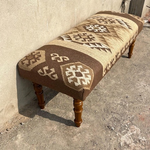 May include: A rectangular bench with a patterned textile top in shades of brown, beige, and white. The bench has four wooden legs with a dark brown finish. The textile features geometric designs.