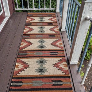 Könnte beinhalten: Ein rechteckiger, gewebter Läuferteppich mit einem südwestlichen Design. Der Teppich weist ein sich wiederholendes Muster aus geometrischen Formen in Beige-, Rost-, Schwarz- und Grautönen auf. Der Teppich hat einen Fransenrand und liegt auf einer Holzveranda.