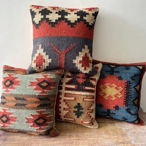 Puede incluir: Cuatro cojines decorativos con patrones geométricos en tonos rojos, azules, grises y beige. Los cojines son cuadrados y parecen estar hechos de tela tejida. Están colocados sobre una superficie de madera.