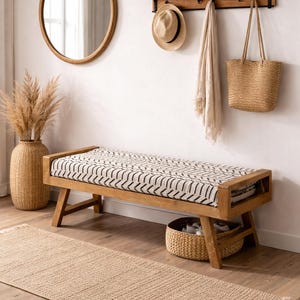 Può includere: Una panca in legno con cuscino fantasia, uno specchio rotondo e un tappeto tessuto. Una mensola con ganci tiene un cappello, un asciugamano e una borsa tote. Un vaso tessuto contiene piante essiccate. La stanza ha una palette di colori neutri.
