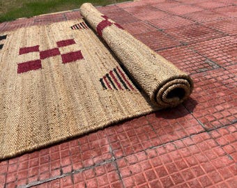 May include: A natural fiber rug with a rolled section. The rug features a woven design with geometric patterns in black and burgundy. The rug is partially rolled up, revealing its texture and construction. The rug is on a red brick surface.