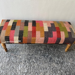 May include: A rectangular bench with a colorful patchwork design. The seat features a pattern of various rectangular shapes in shades of red, pink, orange, green, gray, and black. The bench has four wooden legs.