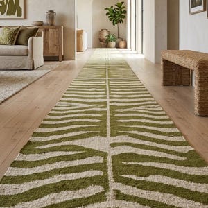 Puede incluir: Una alfombra larga y rectangular con un patrón de rayas verdes y blancas, que se asemeja a un estampado de cebra, está colocada sobre un suelo de madera. La alfombra tiene flecos en el extremo. La alfombra está en un pasillo.