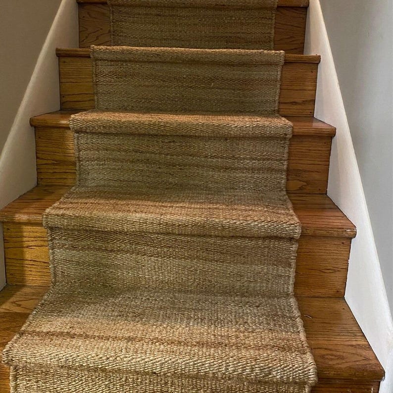 May include: A set of natural fiber stair treads in a light brown color. The treads are woven and have a subtle striped pattern. They are placed on wooden stairs.